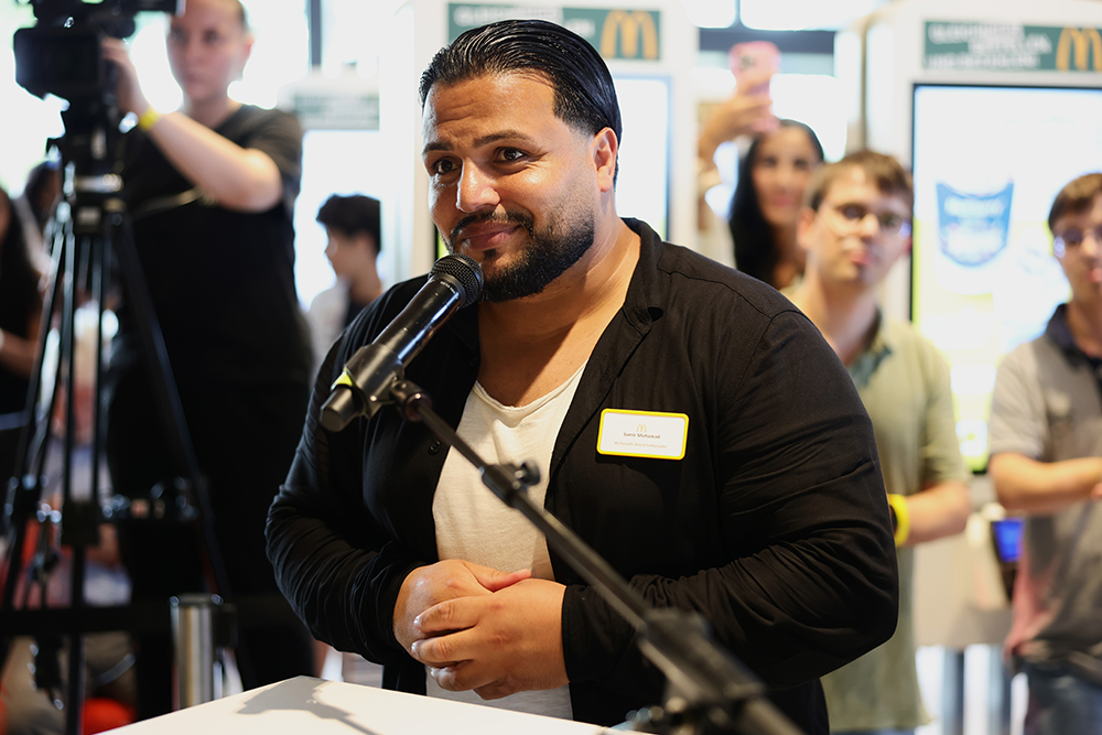 Restaurantmanager Samir Mohamad spricht beim Burger Dialog in Köln-Kalk am Mikrofon, im Hintergrund Gäste und ein Kamerateam.