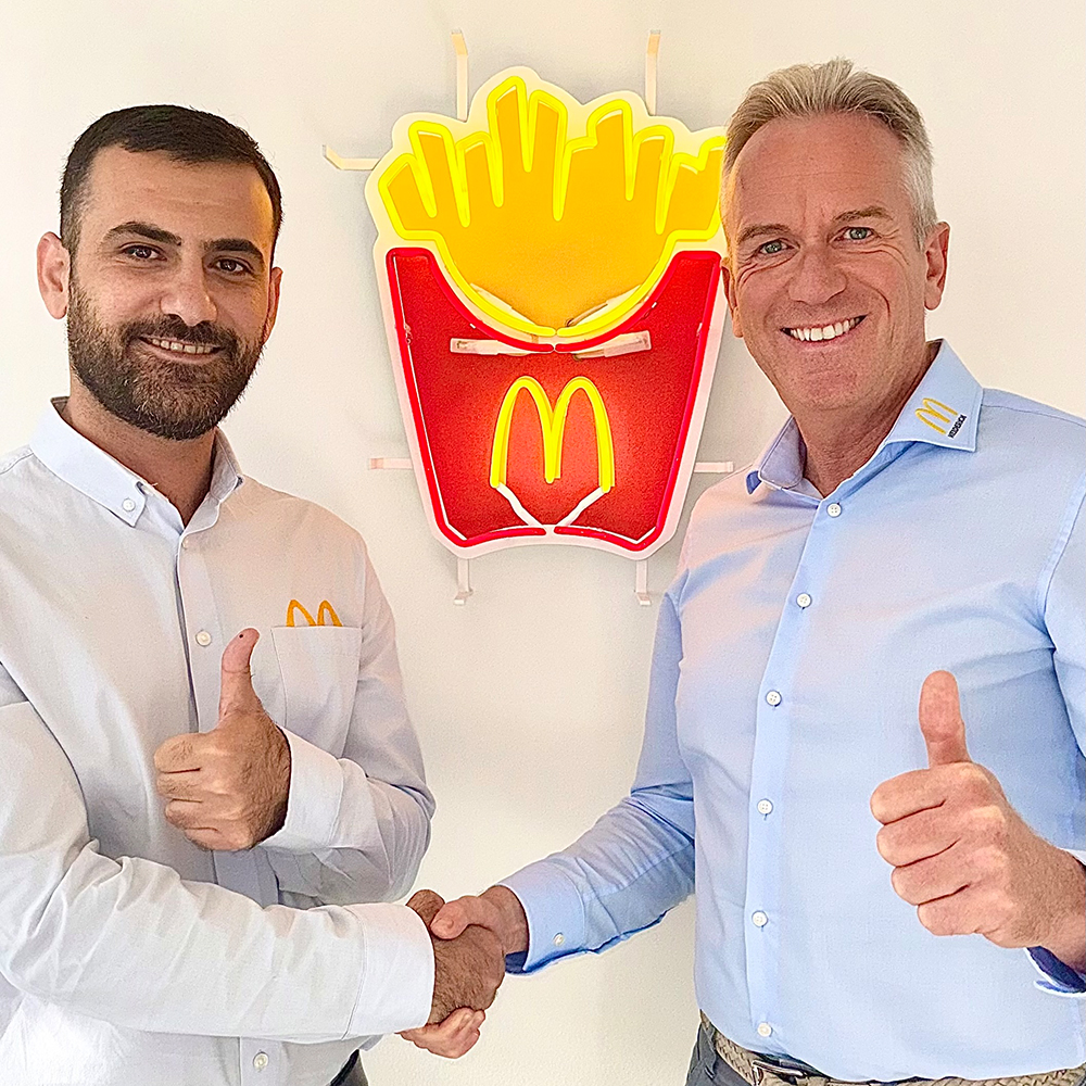 Operations Manager Odah Aladioui und McDonald's Franchise-Nehmer Arndt Heiderich beim Handschlag vor einer Pommes-Leuchtreklame mit McDonald's-Logo